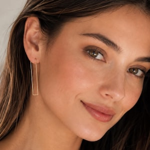 May include: Gold-colored rectangular drop earrings. The earrings are a simple, minimalist design, with a long, slender rectangular shape. The earrings are hanging from the ear. The earrings are a warm gold color.