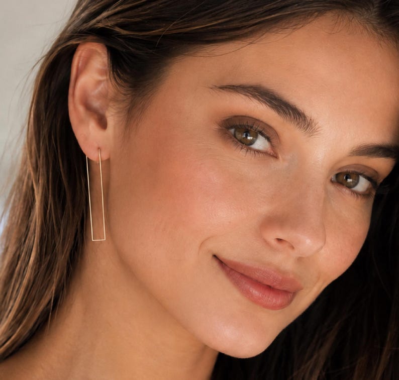 May include: Gold-colored rectangular drop earrings. The earrings are a simple, minimalist design, with a long, slender rectangular shape. The earrings are hanging from the ear. The earrings are a warm gold color.