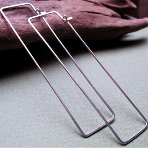 May include: A pair of silver-toned, rectangular hoop earrings. The earrings are long and slender, with a simple, minimalist design. The rectangular shape is formed by thin, straight lines, creating a modern and geometric look.
