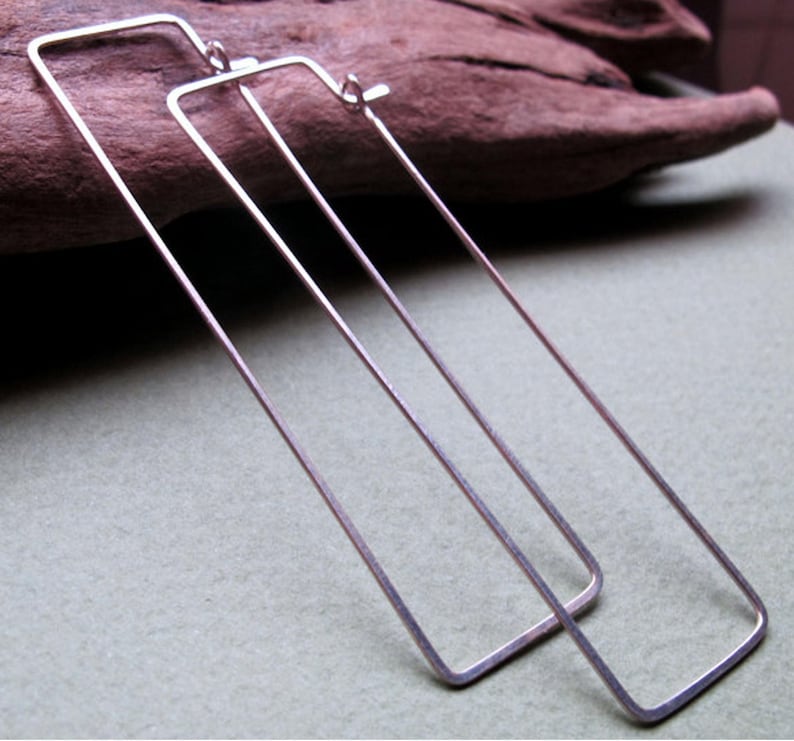 May include: A pair of silver-toned, rectangular hoop earrings. The earrings are long and slender, with a simple, minimalist design. The rectangular shape is formed by thin, straight lines, creating a modern and geometric look.