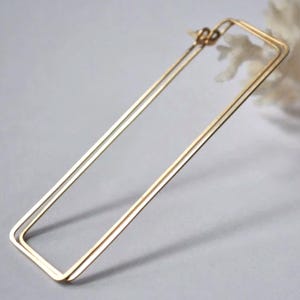 Könnte beinhalten: Goldene rechteckige Creolen. Die Ohrringe bestehen aus zwei dünnen Metallstücken, die zu einem Rechteck zusammengefügt sind.