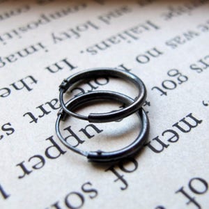 May include: Two silver hoop earrings, one slightly larger than the other, are lying on a page of a book. The earrings are made of a smooth metal and have a simple, classic design.