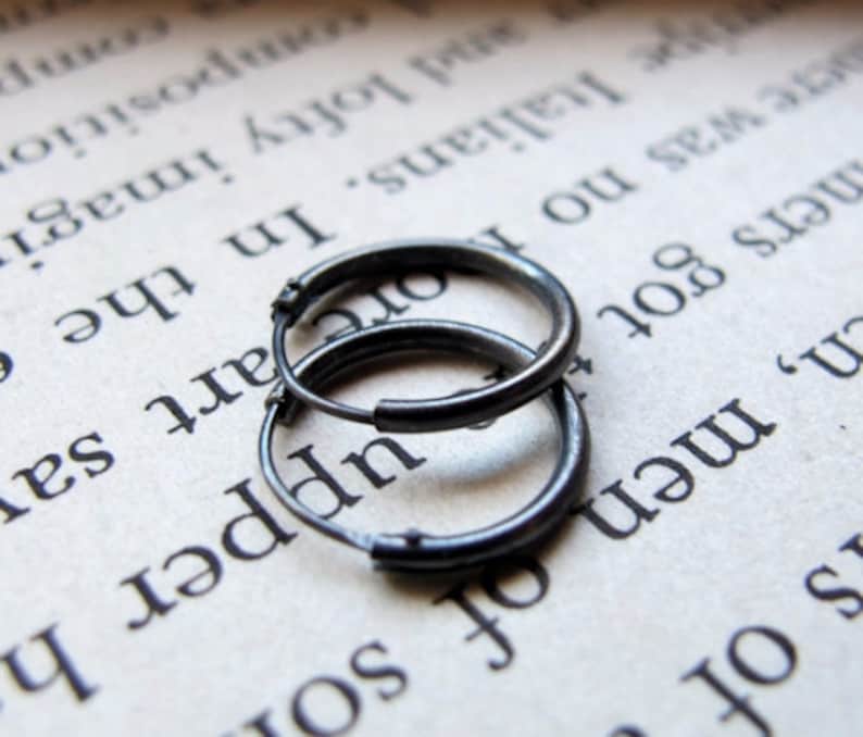 May include: Two silver hoop earrings, one slightly larger than the other, are lying on a page of a book. The earrings are made of a smooth metal and have a simple, classic design.