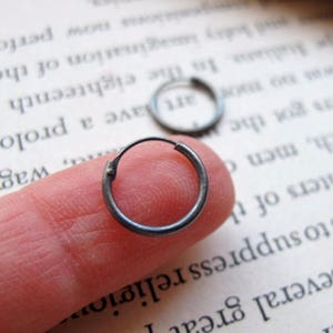May include: A pair of small, silver hoop earrings. The earrings are shown on a finger, with one earring in focus.