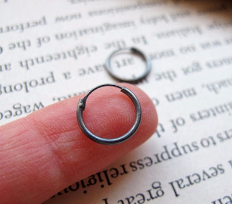 May include: A pair of small, silver hoop earrings. The earrings are shown on a finger, with one earring in focus.