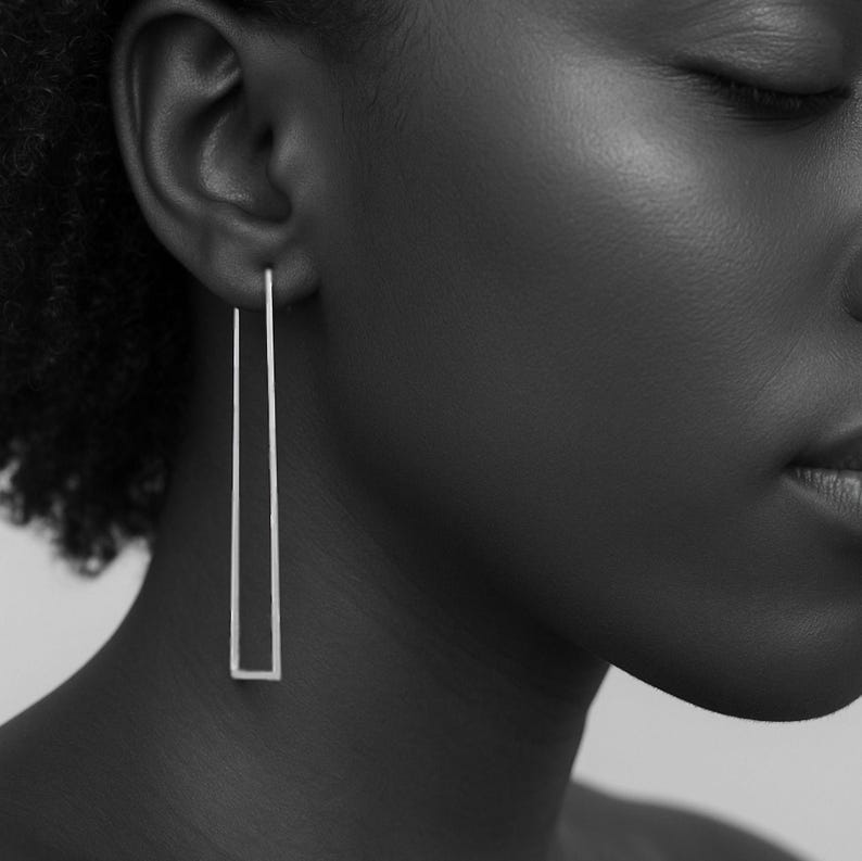 May include: Silver rectangular drop earrings. The earrings are long, thin, and straight, with a minimalist design. The earrings are hanging from the earlobe. The image is in black and white.