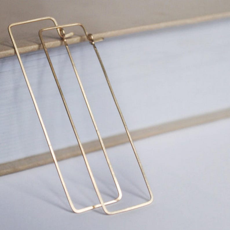 May include: A pair of gold-colored rectangular hoop earrings. The earrings have a simple, minimalist design with a long, slender rectangular shape. The earrings are made of a shiny, metallic material.