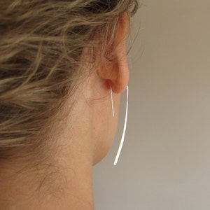 May include: A pair of silver dangle earrings with a thin, long, curved design. The earrings are worn in a person's ear.