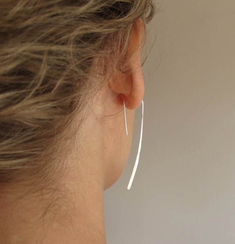 May include: A pair of silver dangle earrings with a thin, long, curved design. The earrings are worn in a person's ear.