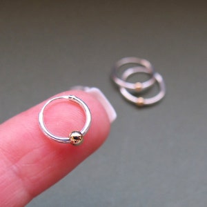 May include: Three silver hoop earrings with a gold bead accent. The earrings are shown on a finger.