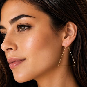 May include: A thin, gold-toned metal earring shaped like an equilateral triangle. The earring features a simple wire frame design with a small hinge mechanism at the top corner for closure, worn in the earlobe as a geometric piece of jewelry.