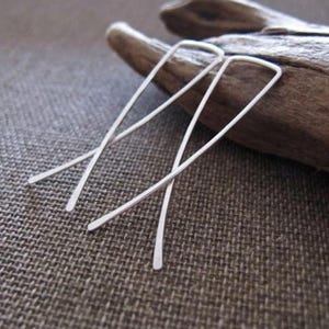 Könnte beinhalten: Ein Paar silberfarbene, minimalistische Ohrringe. Jeder Ohrring hat einen geraden, dünnen Stab, der sich oben krümmt und unten spitz zuläuft. Die Ohrringe werden auf einem Stück Treibholz vor einem strukturierten, neutralfarbenen Hintergrund präsentiert.