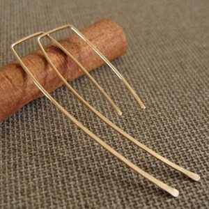 May include: A pair of gold-toned earrings with a simple, rectangular design. The earrings are long and slender, with a slightly curved shape.