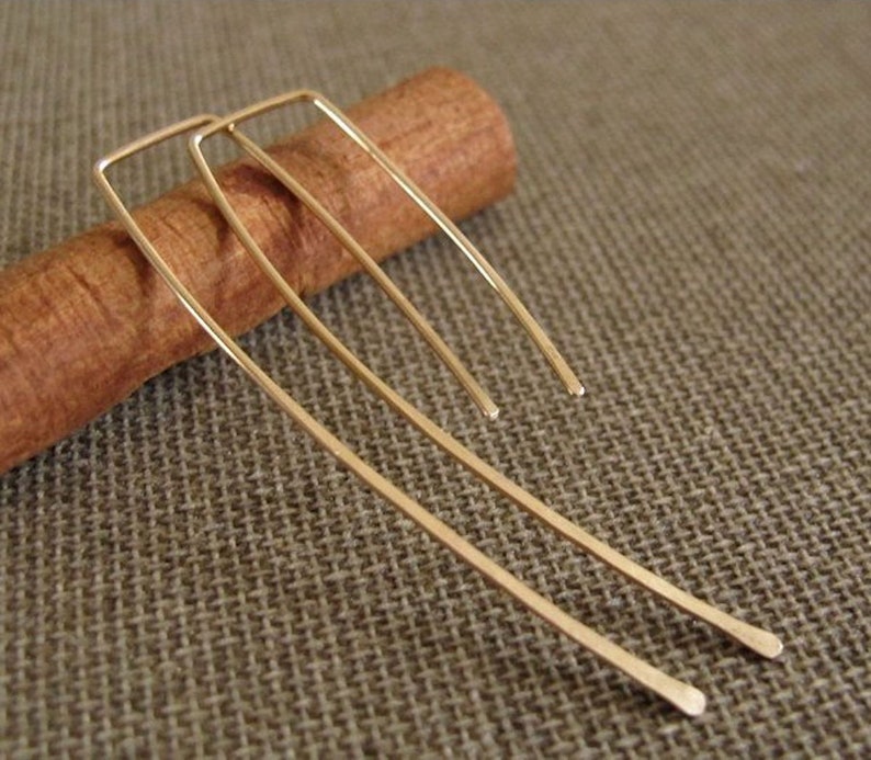 May include: A pair of gold-toned earrings with a simple, rectangular design. The earrings are long and slender, with a slightly curved shape.