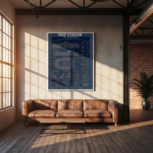 May include: A framed navy blue poster of Ohio Stadium, 'The Horseshoe', with white architectural diagrams and text. The poster hangs above a brown leather sofa and wooden coffee table in a modern living room. The room has wooden flooring.