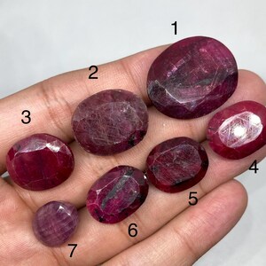 May include: Seven faceted rubies of varying sizes and shapes, including oval and round cuts. The gemstones are a deep red colour, with visible internal inclusions. The rubies are displayed against a light background.