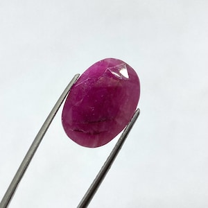 Op de afbeelding: Een gefacetteerde, ovaalvormige robijn edelsteen, vastgehouden door een zilveren pincet. De robijn heeft een diepe, verzadigde roze-rode kleur, met zichtbare interne insluitsels. De edelsteen is gepolijst en reflecteert licht.