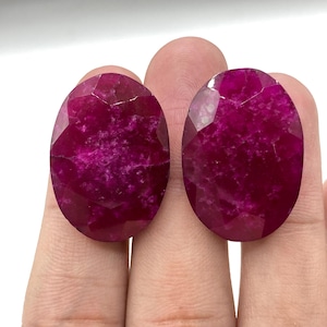 May include: Two oval-shaped, faceted gemstones with a deep magenta color. The gems have a polished surface with visible internal inclusions. The stones are held between fingers, showcasing their size and color.