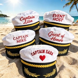 Puede incluir: Cinco gorras de capitán blancas con bordes negros y adornos dorados están dispuestas en una playa de arena. Cada gorra tiene texto bordado en rojo: "Captain Lisa", "Event Host", "Captain Mike", "Guest of Honor" y "Captain Sara" con un corazón rojo.