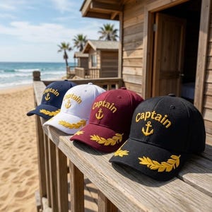 May include: Four baseball caps in navy, white, burgundy, and black, each with "captain" embroidered in gold, along with an anchor and laurel leaf design. The caps are displayed on a wooden railing with a beach and ocean backdrop.