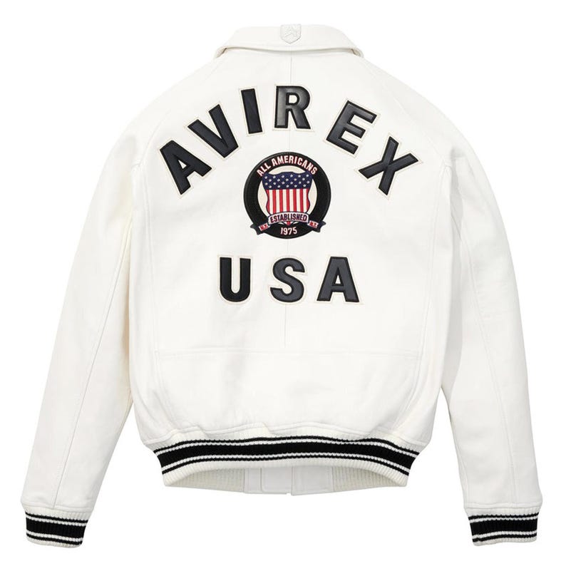 Peut inclure: Veste en cuir blanche avec des poignets et un ourlet ray&eacute;s noirs et blancs. Le dos pr&eacute;sente le texte "AVIREX" au-dessus d'un embl&egrave;me circulaire avec un drapeau am&eacute;ricain et le mot "USA".