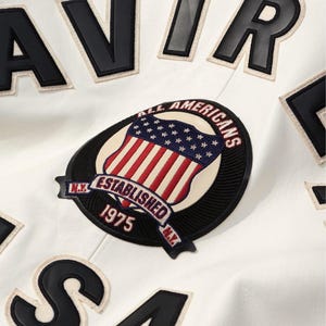 Peut inclure: Gros plan sur une veste blanche avec des lettres et des &eacute;cussons noirs. La veste pr&eacute;sente un &eacute;cusson avec un drapeau am&eacute;ricain et le texte "ALL AMERICANS ESTABLISHED 1975 N.Y."