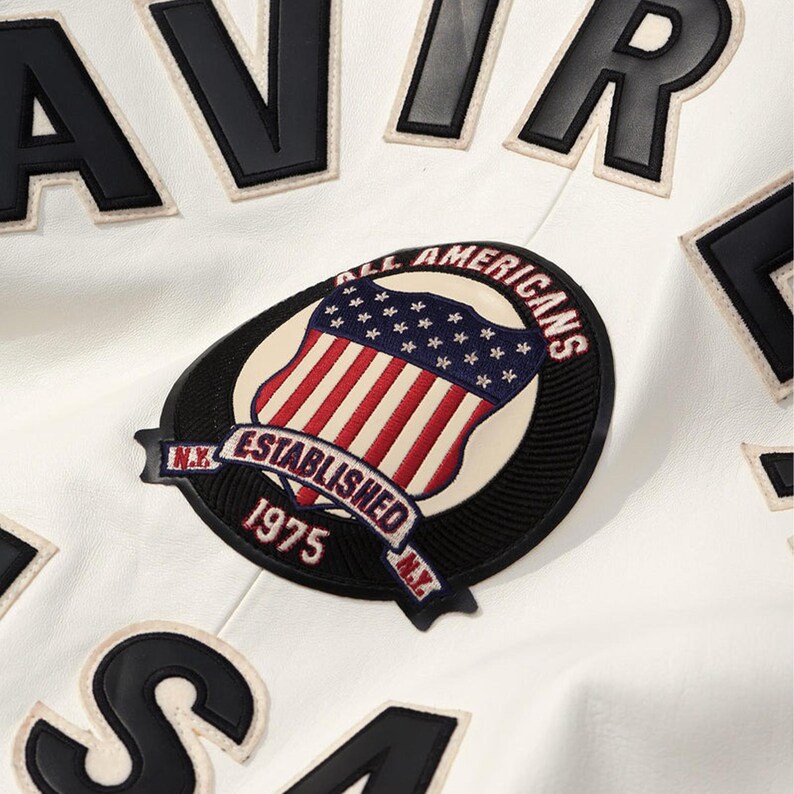 Peut inclure: Gros plan sur une veste blanche avec des lettres et des &eacute;cussons noirs. La veste pr&eacute;sente un &eacute;cusson avec un drapeau am&eacute;ricain et le texte "ALL AMERICANS ESTABLISHED 1975 N.Y."