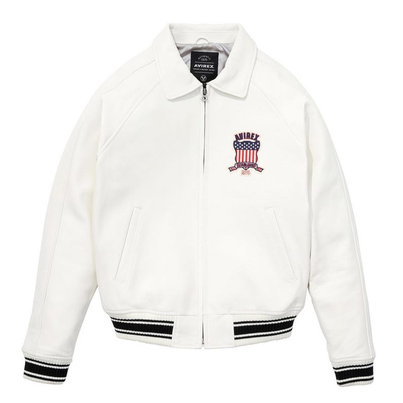 Peut inclure: Veste de bombardier blanche avec col et fermeture &eacute;clair. Le blouson pr&eacute;sente des poignets et un ourlet ray&eacute;s noirs et blancs. Un &eacute;cusson brod&eacute; avec le texte "AVIREX" et un motif de drapeau am&eacute;ricain est sur la poitrine.