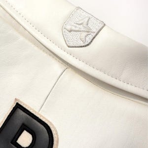 Peut inclure: Gros plan sur une veste en cuir blanche avec une couture et deux &eacute;cussons. L'un est un embl&egrave;me brod&eacute; blanc, l'autre est une lettre "D" noire et blanche. La veste a une texture lisse.