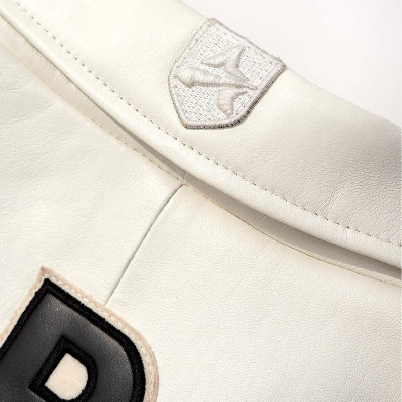 Peut inclure: Gros plan sur une veste en cuir blanche avec une couture et deux &eacute;cussons. L'un est un embl&egrave;me brod&eacute; blanc, l'autre est une lettre "D" noire et blanche. La veste a une texture lisse.