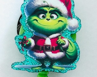 Puede incluir: Un portacredenciales con un borde azul cerceta brillante y una ilustración impresa de un personaje verde con un traje y gorro de Papá Noel rojo y blanco, sosteniendo un pequeño regalo y un saco verde, unido a un clip de plástico verde lima.