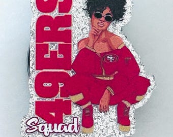 Può includere: Un portabadge glitterato con la grafica di una persona con capelli scuri ricci, occhiali da sole e un completo rosso con il logo 49ers. Il testo 49ERS è stampato in grandi lettere rosse verticalmente, con la parola Squad in corsivo.