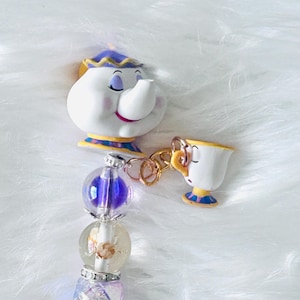 Peut inclure: Un stylo décoratif avec un embout en forme de théière blanche aux accents violets et dorés, une petite breloque tasse attachée par un anneau doré, et des perles dont une sphère violette translucide, une perle florale et une perle bleue scintillante.