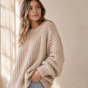 Könnte beinhalten: Ein beigefarbener Grobstrickpullover mit geripptem Rundhalsausschnitt, langen Ärmeln mit gerippten Bündchen und einem vertikalen Strukturmuster. Der Pullover hat eine lockere, übergroße Passform und wird mit einer hellblauen Jeans kombiniert.