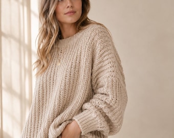 Puede incluir: Un suéter de punto grueso en color beige con cuello redondo acanalado, mangas largas con puños acanalados y un patrón de textura vertical. El suéter presenta un corte holgado y extragrande, combinado con pantalones vaqueros azul claro.