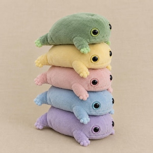 May include: A vertical stack of five plush frog toys in soft pastel colors. From top to bottom, the frogs are sage green, pale yellow, light pink, sky blue, and lavender. Each frog features a rounded body, small limbs, and large black button eyes.
