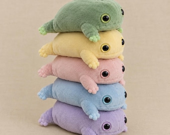 Puede incluir: Una pila vertical de cinco juguetes de peluche con forma de rana en colores pastel suaves. De arriba a abajo, las ranas son verde salvia, amarillo pálido, rosa claro, azul cielo y lavanda. Cada rana tiene un cuerpo redondeado y ojos negros.