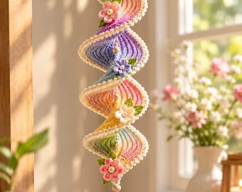 Puede incluir: Un adorno de ganchillo en espiral colgante con un degradado de colores pastel que incluye rosa, morado, azul, amarillo y verde. La pieza está decorada con pequeñas flores de ganchillo, hojas verdes y un cristal facetado transparente.