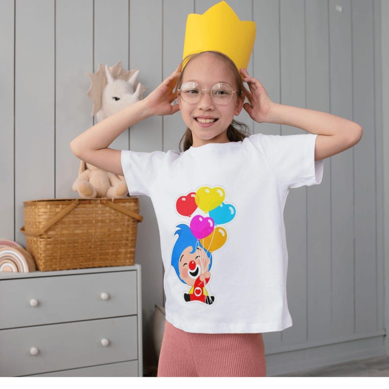 May include: A white t-shirt featuring a cartoon character holding heart-shaped balloons in red, yellow, pink, and blue. The person is wearing a yellow paper crown and glasses. The background includes a gray dresser and a wicker basket.