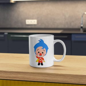 May include: White ceramic mug featuring a cartoon character with blue hair, a red nose, and a red and yellow outfit. The character is waving. The mug has a curved handle and is set on a wooden surface.