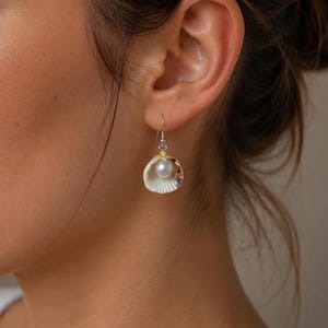 May include: A drop earring featuring a small, off-white seashell with a smooth, lustrous white pearl nestled inside the shell cavity. The shell is suspended from a thin, silver-coloured metal hook wire that passes through the earlobe.