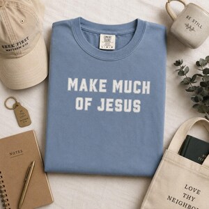 May include: A blue crew neck t-shirt with the white text "MAKE MUCH OF JESUS" printed on the front. The shirt features a Comfort Colors brand label at the inner neckline. The garment is displayed alongside a tan baseball cap, a notebook, a pen, a mug, and a tote bag.