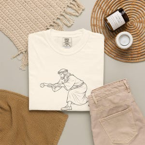 Könnte beinhalten: Ein cremefarbenes T-Shirt mit einer schwarzen Strichzeichnung einer Figur mit Dornenkrone, die einen Baseballschläger in Schlagposition hält. Das T-Shirt hat ein Comfort Colors Markenetikett am inneren Halsausschnitt mit der aufgedruckten Größe L G G.