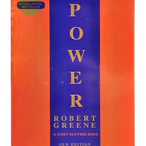 May include: A book titled "The Concise 48 Laws of Power" by Robert Greene. The cover features a vertical blue stripe with the word "POWER" in large yellow letters. The book has an orange background and an "INTERNATIONAL BESTSELLER" sticker.