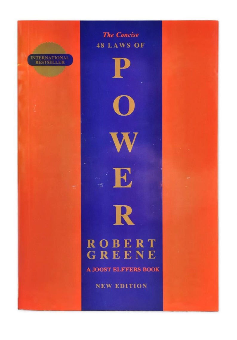 May include: A book titled "The Concise 48 Laws of Power" by Robert Greene. The cover features a vertical blue stripe with the word "POWER" in large yellow letters. The book has an orange background and an "INTERNATIONAL BESTSELLER" sticker.