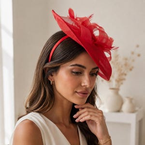 Könnte beinhalten: Ein leuchtend roter Fascinator mit einer breiten, kreisförmigen Krempe und komplexen, geschichteten Netzschleifen. Das Kopfstück ist mit einem passenden roten Haarreif befestigt und zeigt zarte Federakzente, die dem Design Struktur verleihen.