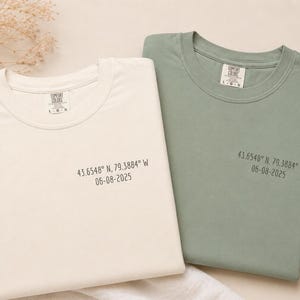 以下が含まれることがあります： クリーム色とセージグリーンの2枚の折り畳まれたTシャツが表示されています。各シャツの前面には「43.6548° N, 79.3884° W 06-08-2025」の文字がプリントされています。シャツは白い表面にあります。