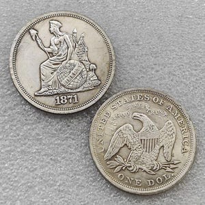 May include: Two silver-colored coins. One coin shows a seated figure holding a pole with a cap, resting a hand on a globe labeled LIBERTY, with the year 1871. The other coin features an eagle with a shield, the text UNITED STATES OF AMERICA, IN GOD WE TRUST, and ONE DOL.