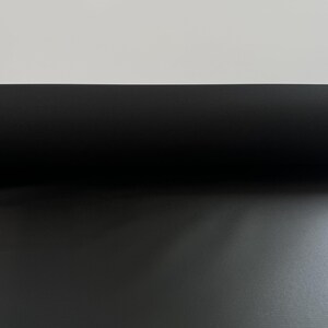 May include: A roll of solid black fabric with a fine, textured weave. The material is tightly wound, creating a smooth, cylindrical shape that transitions from a dark, shadowed upper section to a lighter, textured lower surface.