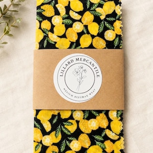 May include: A stack of rectangular beeswax wraps featuring a black background with a yellow lemon and green leaf pattern. A brown paper band wraps around the center with a circular white label reading Lillard Mercantile Medium Beeswax Wrap.